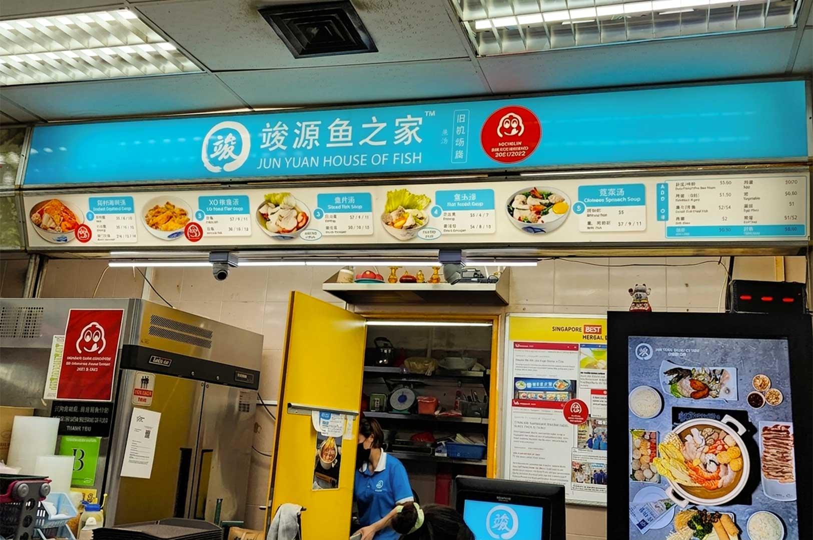 A eye-level view of the Jun Yuan House of Fish stall counter, detailing an overhead bright teal signboard displaying menu options with prices alongside photos of dishes, kitchen shelves visible through a yellow-framed doorway, and framed news articles celebrating the stall's awards.