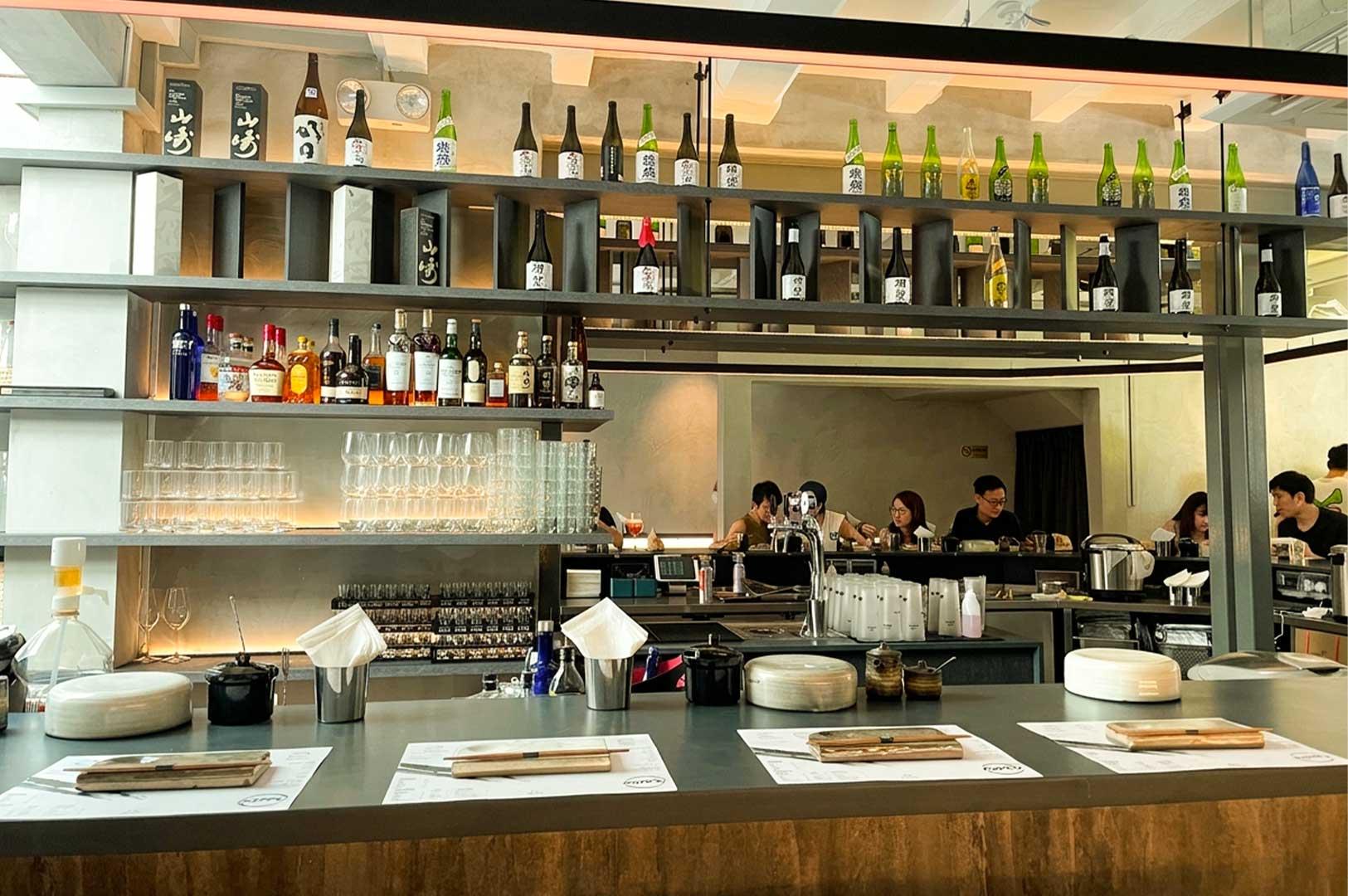 A modern Japanese handroll bar featuring a long dining counter set with individual placemats, wooden blocks holding chopsticks, and white paper napkins. Overhead, multi-tiered shelving holds a vast collection of sake and liquor bottles along with stacked glassware. In the background, patrons sit across the central bar area, enjoying their meals in a warm, warmly lit interior.