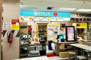 The storefront of Jun Yuan House of Fish at a food center, featuring a teal and white sign with Chinese characters and English text, a menu board displaying various fish soup options above an open kitchen area, an illuminated menu display near the POS counter, and stainless steel dining tables with stools nearby.