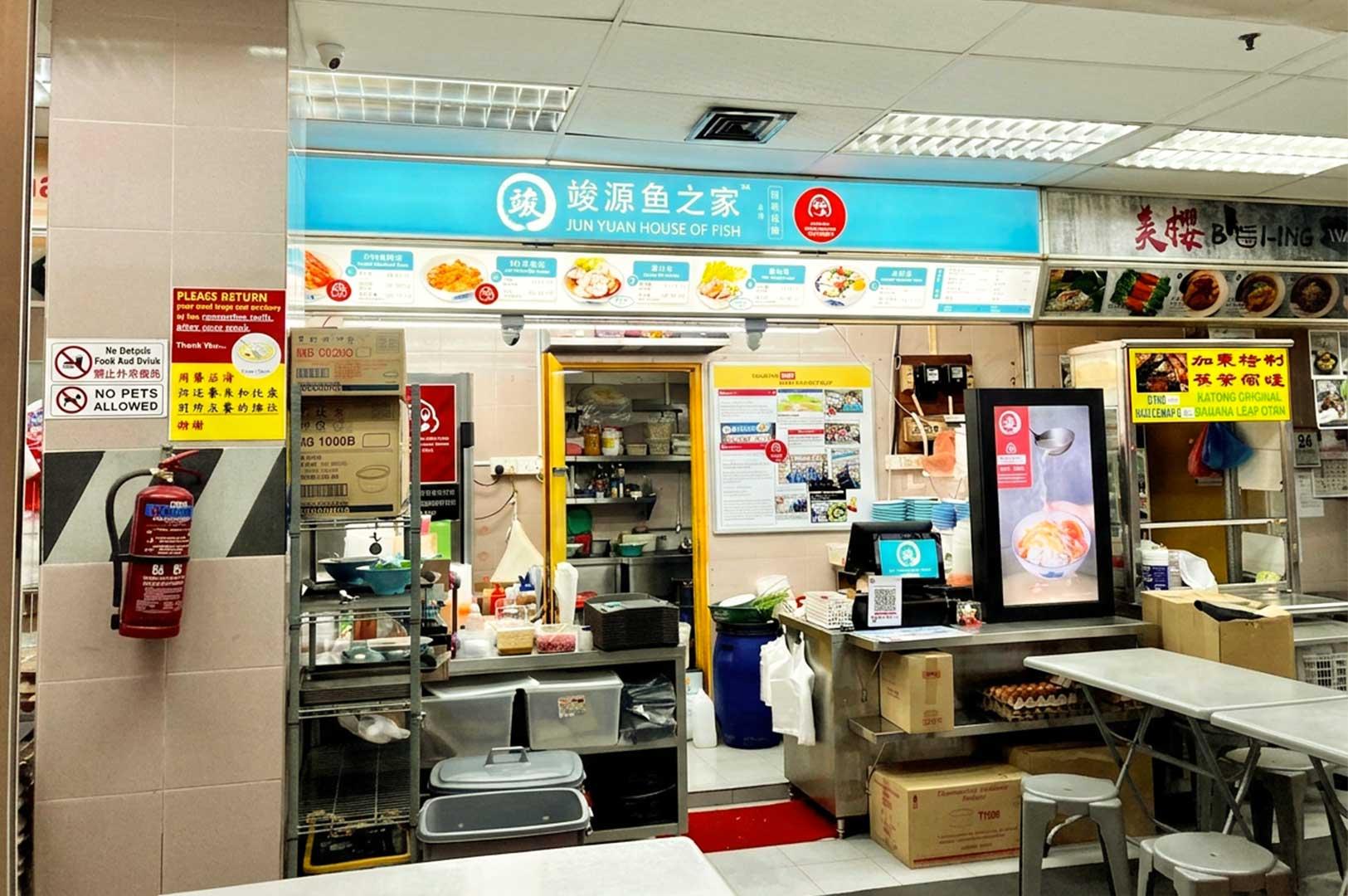 The storefront of Jun Yuan House of Fish at a food center, featuring a teal and white sign with Chinese characters and English text, a menu board displaying various fish soup options above an open kitchen area, an illuminated menu display near the POS counter, and stainless steel dining tables with stools nearby.